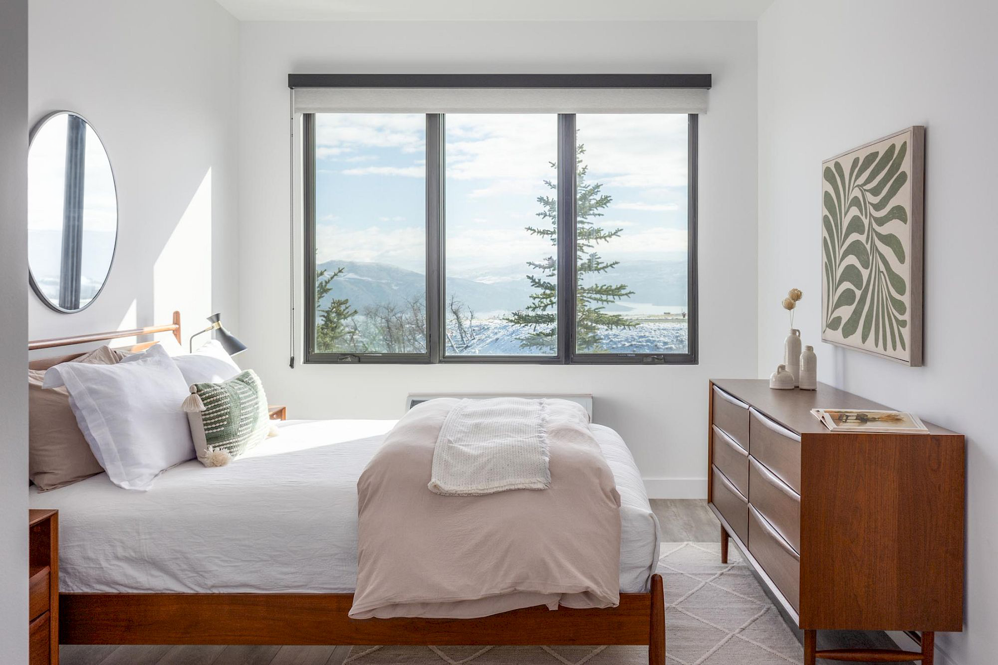 A neatly made bed with pillows and a blanket in a room with a large window, a dresser, and artwork on the wall.
